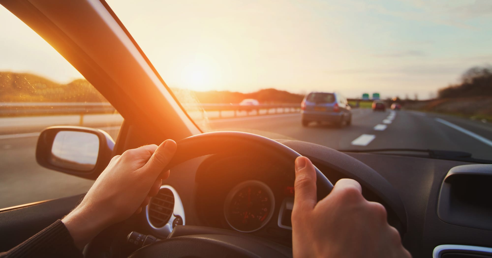 Mãos de homem em volante, na estrada em viagem de carro, com luz do pôr do sol.